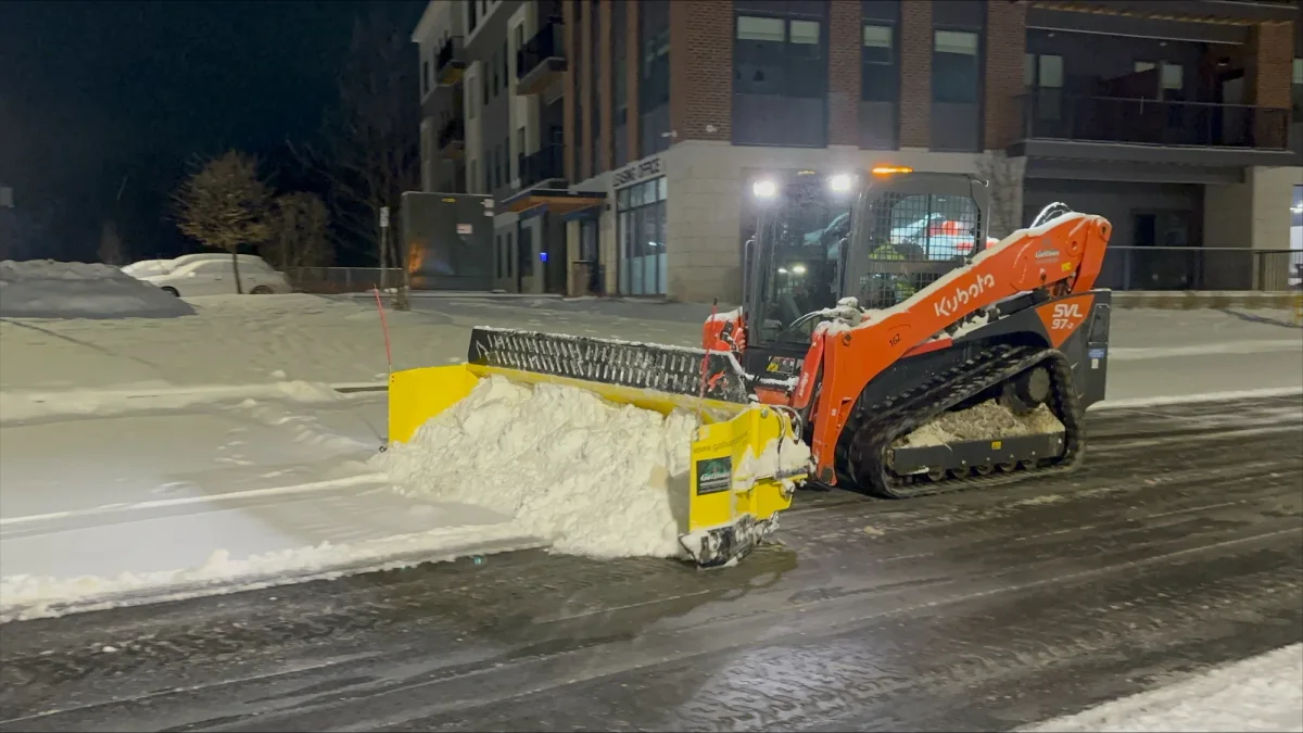 Orange tracked skid steer with a yellow snow pusher clearing snow beside a building at night